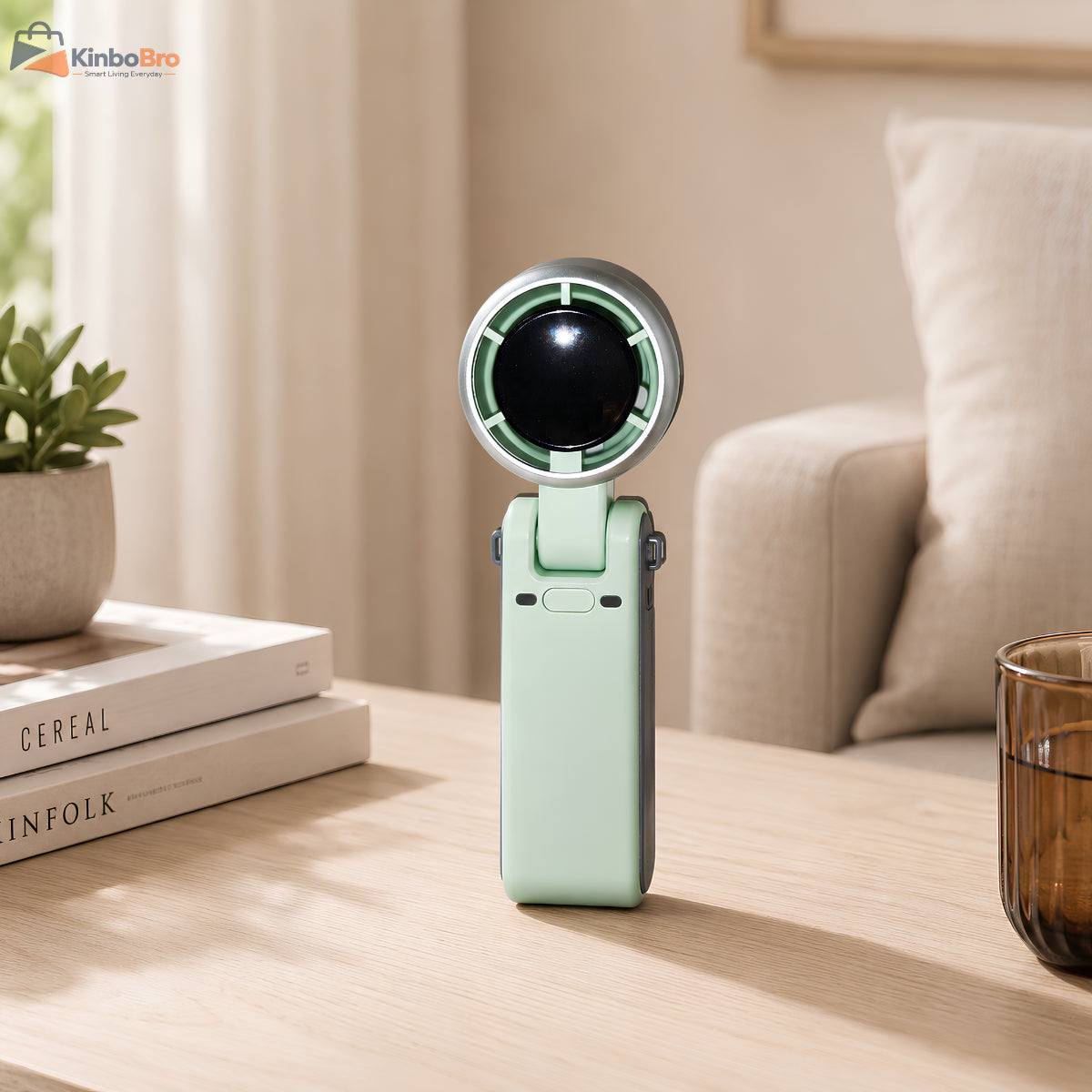 A compact handheld fan in mint green and gray stands on a light wood table in a cozy living room setting, with books, a plant, and a sofa in the background.